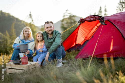 Portrait of a couple with little girl during picnic at campsite while traveling with tent in the mountains. Young family happily spending vacations on nature