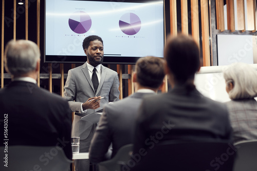 Content confident young Afro-American financial specialist in gray suit standing against plasma screen with graph charts and giving speech in front of business colleagues