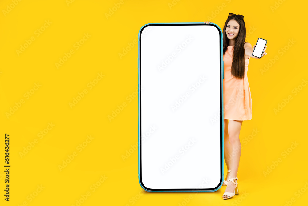 Pretty young asian woman standing near the big smartphone with mockup white screen for your app, text or advertisement design. Blank cellphone in her hand. Studio shoot over the yellow background. Pretty young asian woman standing near the big smartphone with mockup white screen for your app, text or advertisement design. Blank cellphone in her hand. Studio shoot over the yellow background.