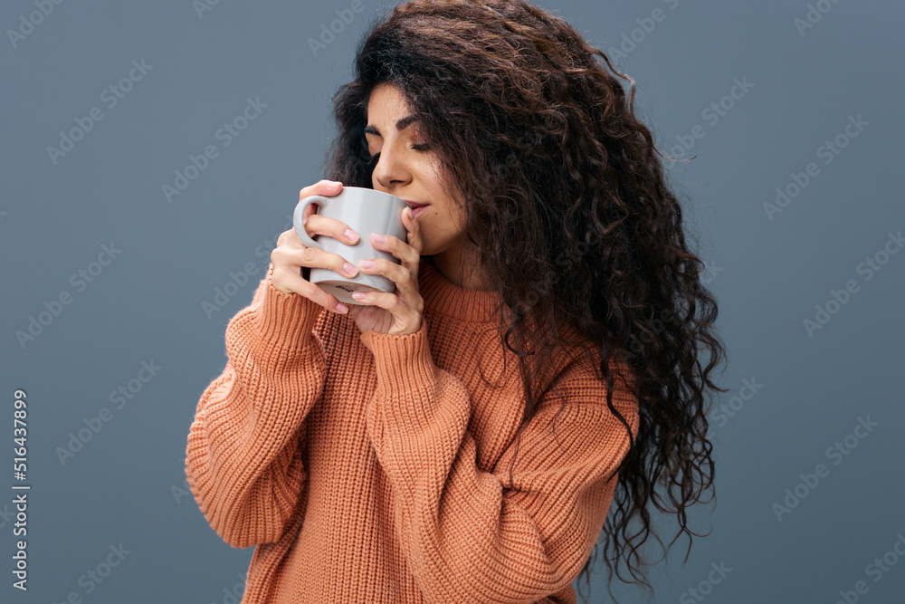 COFFEE TIME CONCEPT. Enjoyed tanned curly Latin female in warm sweater holding cup of tea by both hands posing isolated over gray blue background close eyes. Copy space Mockup Banner. Fashion offer COFFEE TIME CONCEPT. Enjoyed tanned curly Latin female in warm sweater holding cup of tea by both hands posing isolated over gray blue background close eyes. Copy space Mockup Banner. Fashion offer