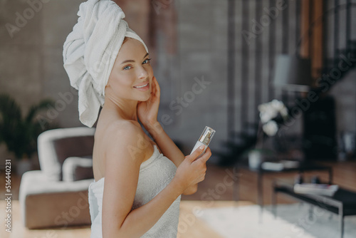 Indoor shot of pretty European woman with healthy glowing skin, minimal makeup, holds bottle of body lotion, pampers complexion, wears bath towel around body, poses against domestic interior
