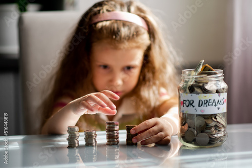 A sweet girl with full piggy bank of coins. On the piggy bank there is an inscription for dream. The girl is at home. She sits at a table and counts the coins. The money. Savings. Childhood.