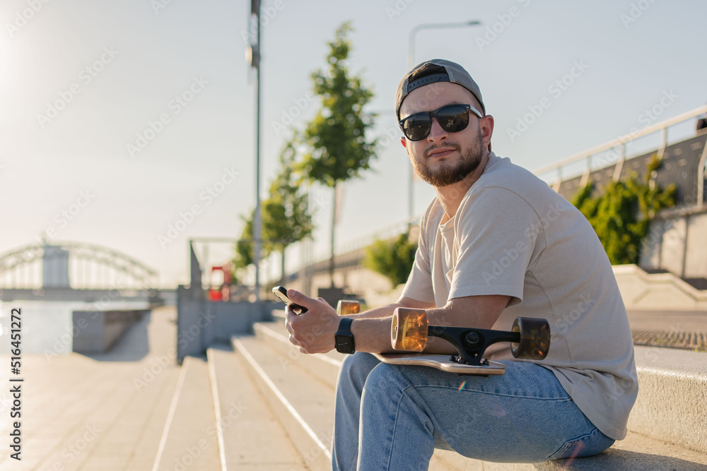 Attractive cheerful young man sitting at the street with a skateboard, using mobile phone.