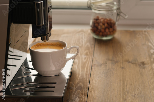 Automatic coffee machine for coffee in white cup near window at daytime in a coffee shop, cafe or restaurant. Jar of hazelnuts on background