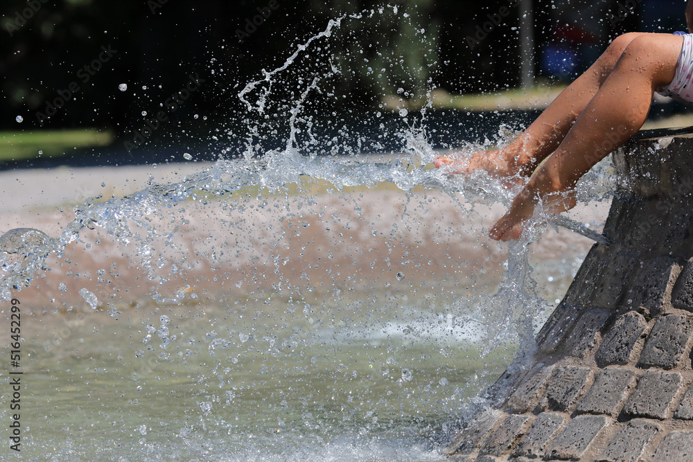 Beautiful legs of a girl sitting on a city fountain. Selective focus. Jets of cool water wash women's feet on a hot summer day.