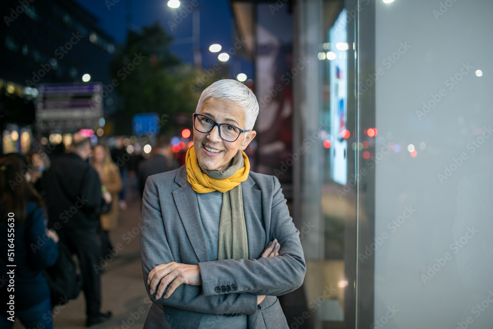 Fototapeta premium Smiling mature senior woman with short gray hair and eyeglasses walking on street, night scene in city