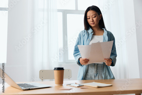 Concentrated thoughtful young Asian cute female businesswoman deals with documents on table with laptop in office interior. Employee work at home. Lady corporation leader concept. Copy space Offer © SHOTPRIME STUDIO