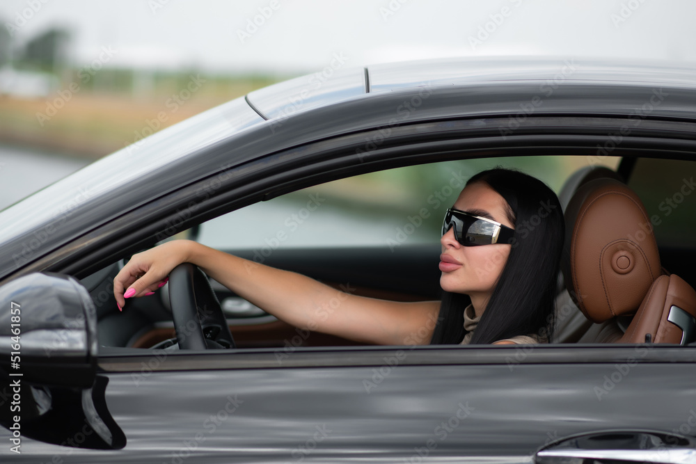Naklejka premium Beautiful brunette in a black cap sits in a black car with her hands on the steering wheel