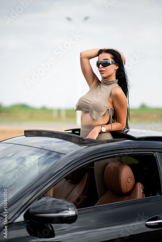 Beautiful brunette in black glasses joyfully leans out of the hatch of a black luxury car. She fixes her hair with her hands
