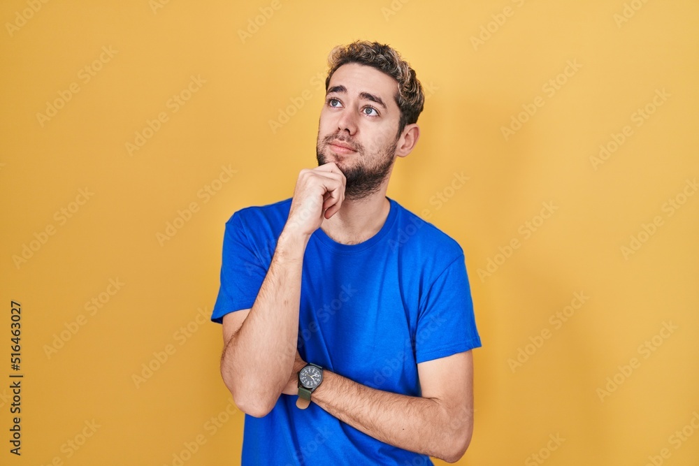 Hispanic man with beard standing over yellow background with hand on chin thinking about question, pensive expression. smiling with thoughtful face. doubt concept. Hispanic man with beard standing over yellow background with hand on chin thinking about question, pensive expression. smiling with thoughtful face. doubt concept.