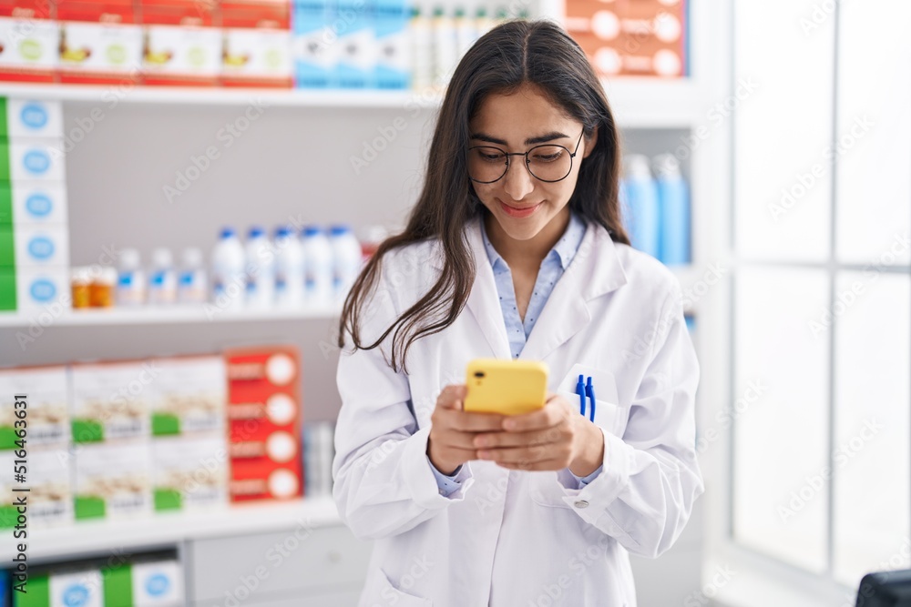 Young hispanic girl pharmacist using smartphone working at pharmacy