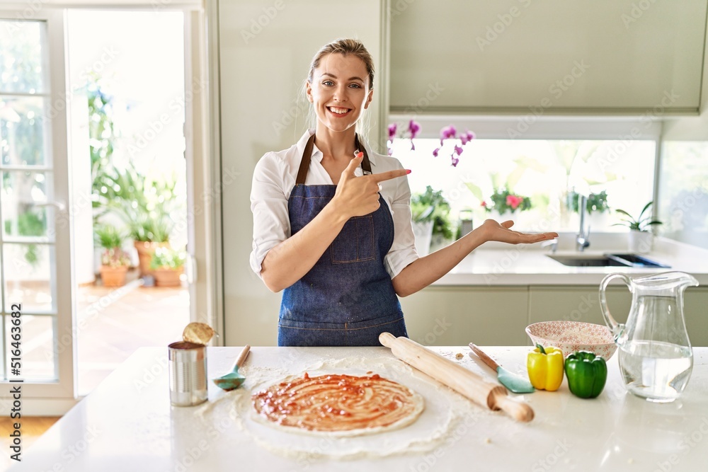 Beautiful blonde woman wearing apron cooking pizza amazed and smiling to the camera while presenting with hand and pointing with finger.