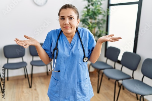 Foto Young brunette doctor woman at waiting room clueless and confused expression with arms and hands raised