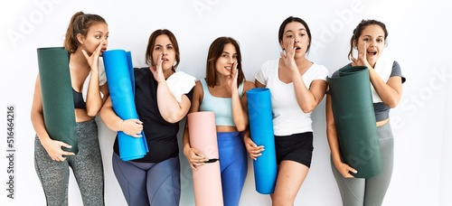 Group of women holding yoga mat standing over isolated background hand on mouth telling secret rumor, whispering malicious talk conversation