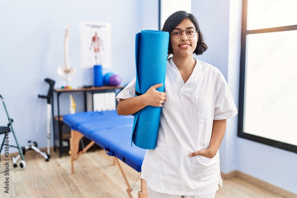 Young latin woman wearing physiotherapist uniform holding yoga mat at physiotherapy clinic