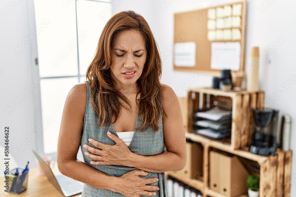 Middle age hispanic woman at the office with hand on stomach because nausea, painful disease feeling unwell. ache concept.