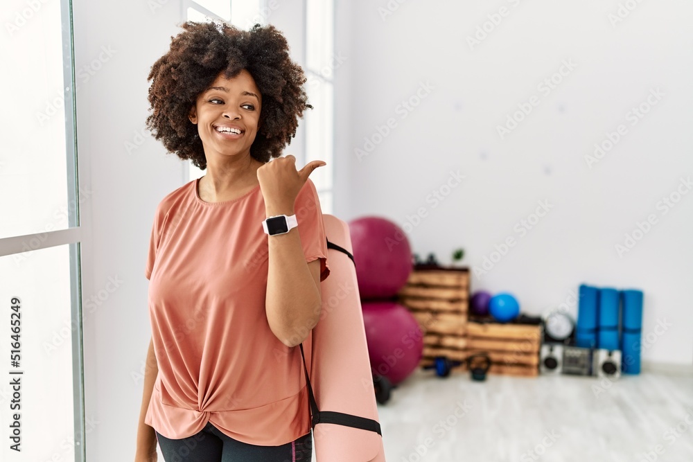 African american woman with afro hair holding yoga mat at pilates room smiling with happy face looking and pointing to the side with thumb up.