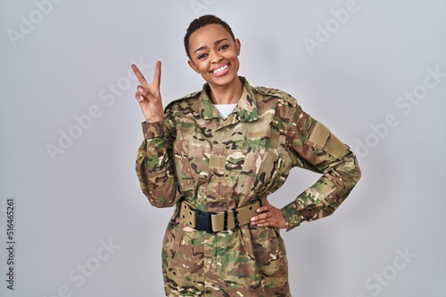 Fotografie Beautiful african american woman wearing camouflage army uniform smiling looking to the camera showing fingers doing victory sign