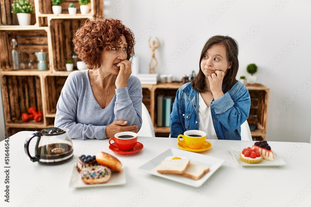 Family of mother and down syndrome daughter sitting at home eating breakfast looking stressed and nervous with hands on mouth biting nails. anxiety problem.