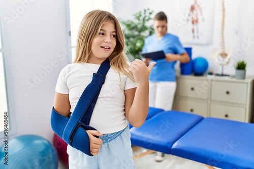 Blonde little girl wearing arm on sling at rehabilitation clinic pointing thumb up to the side smiling happy with open mouth