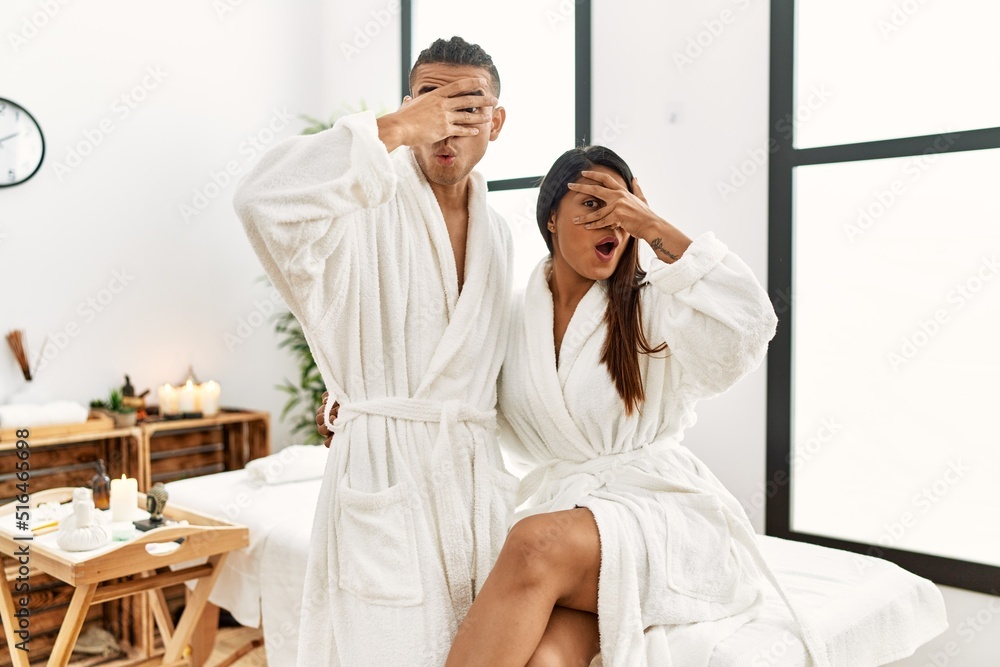 Young latin couple wearing towel standing at beauty center peeking in shock covering face and eyes with hand, looking through fingers with embarrassed expression. Young latin couple wearing towel standing at beauty center peeking in shock covering face and eyes with hand, looking through fingers with embarrassed expression.
