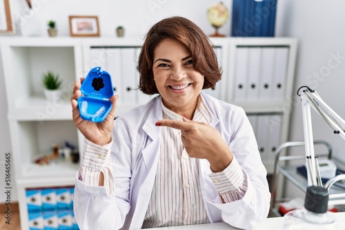 Middle age hispanic dentist woman working at dentist clinic holding invisible aligner smiling happy pointing with hand and finger