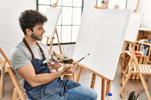 Young hispanic artist man smiling happy holding dog and painting at art studio.