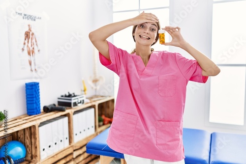 Young physiotherapist woman working at pain recovery clinic holding pills stressed and frustrated with hand on head  surprised and angry face
