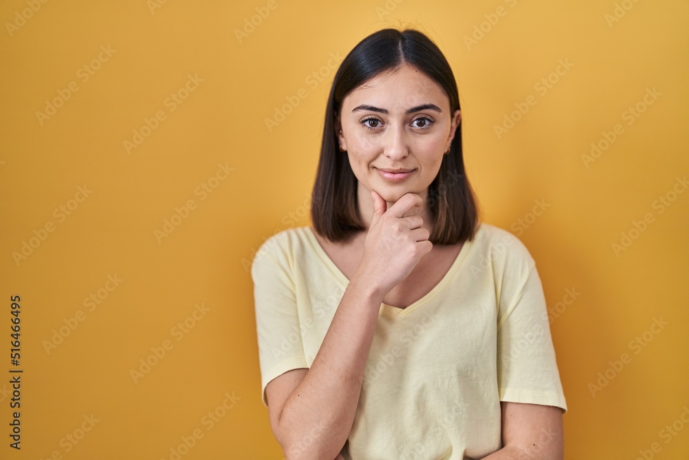 Hispanic girl wearing casual t shirt over yellow background looking confident at the camera smiling with crossed arms and hand raised on chin. thinking positive. Hispanic girl wearing casual t shirt over yellow background looking confident at the camera smiling with crossed arms and hand raised on chin. thinking positive.