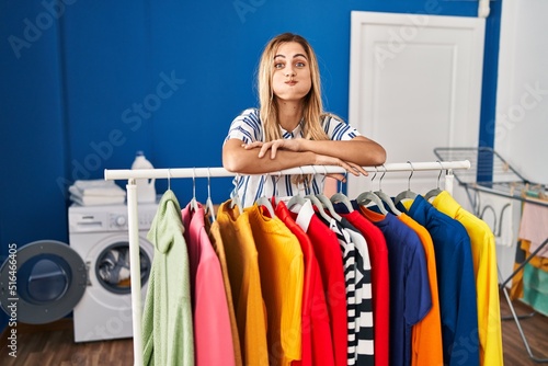 Young blonde woman at laundry room with clean clothes puffing cheeks with funny face. mouth inflated with air, catching air.