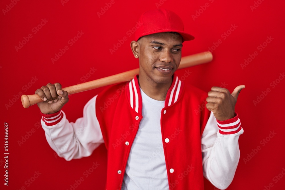 Young hispanic man playing baseball holding bat smiling with happy face looking and pointing to the side with thumb up. Young hispanic man playing baseball holding bat smiling with happy face looking and pointing to the side with thumb up.