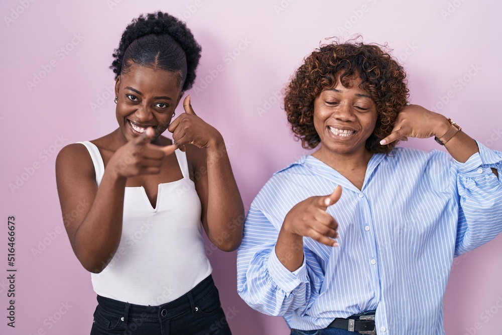 Two african women standing over pink background smiling doing talking on the telephone gesture and pointing to you. call me. Two african women standing over pink background smiling doing talking on the telephone gesture and pointing to you. call me.