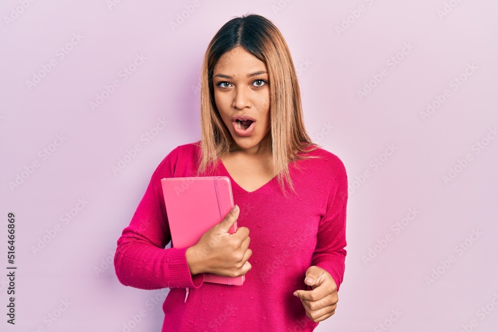 Beautiful hispanic woman holding book scared and amazed with open mouth for surprise, disbelief face Beautiful hispanic woman holding book scared and amazed with open mouth for surprise, disbelief face
