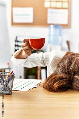 Young hispanic businesswoman overworked with head on table and holding cup of coffee at the office. photo