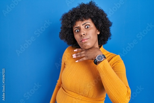 Black woman with curly hair standing over blue background cutting throat with hand as knife, threaten aggression with furious violence