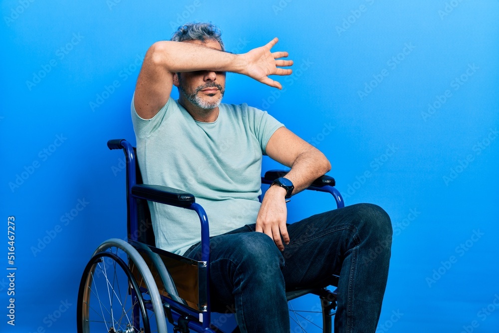 Obraz premium Handsome middle age man with grey hair sitting on wheelchair covering eyes with arm, looking serious and sad. sightless, hiding and rejection concept