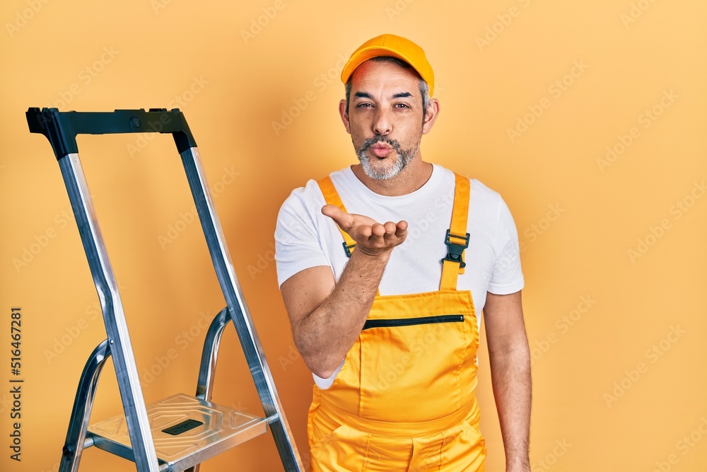 Handsome middle age man with grey hair holding ladder looking at the camera blowing a kiss with hand on air being lovely and sexy. love expression. Handsome middle age man with grey hair holding ladder looking at the camera blowing a kiss with hand on air being lovely and sexy. love expression.