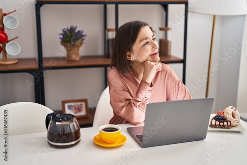 Middle age woman having breakfast using laptop sitting on table at home