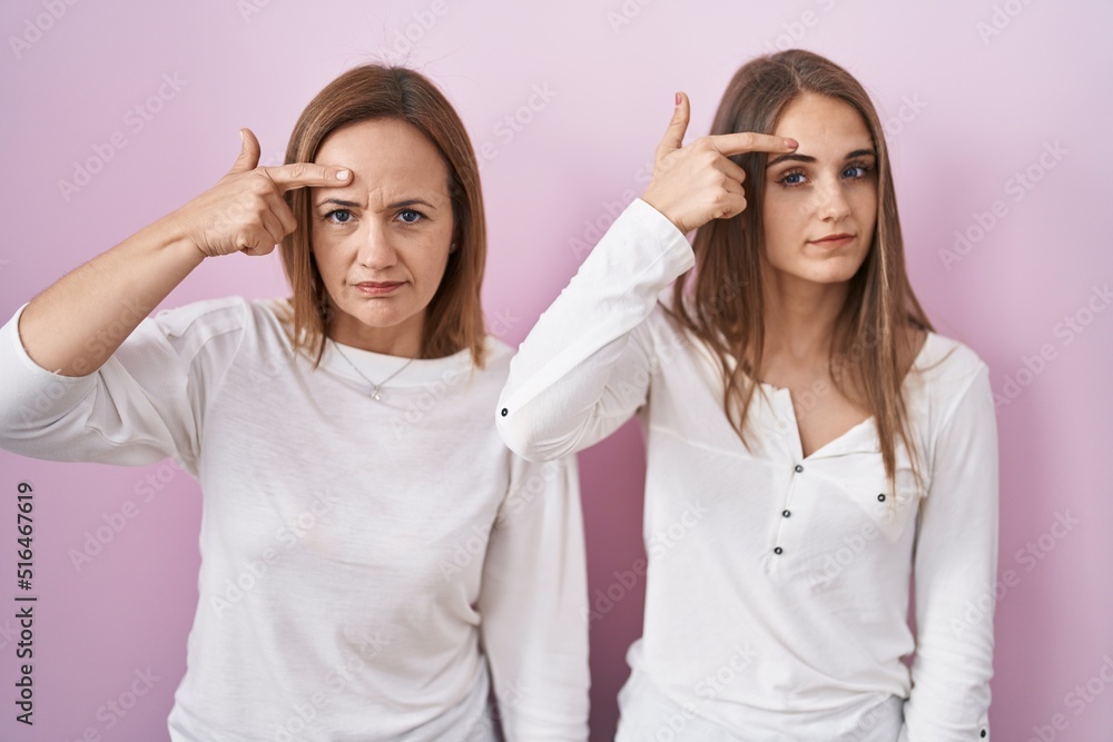Middle age mother and young daughter standing over pink background pointing unhappy to pimple on forehead, ugly infection of blackhead. acne and skin problem Middle age mother and young daughter standing over pink background pointing unhappy to pimple on forehead, ugly infection of blackhead. acne and skin problem