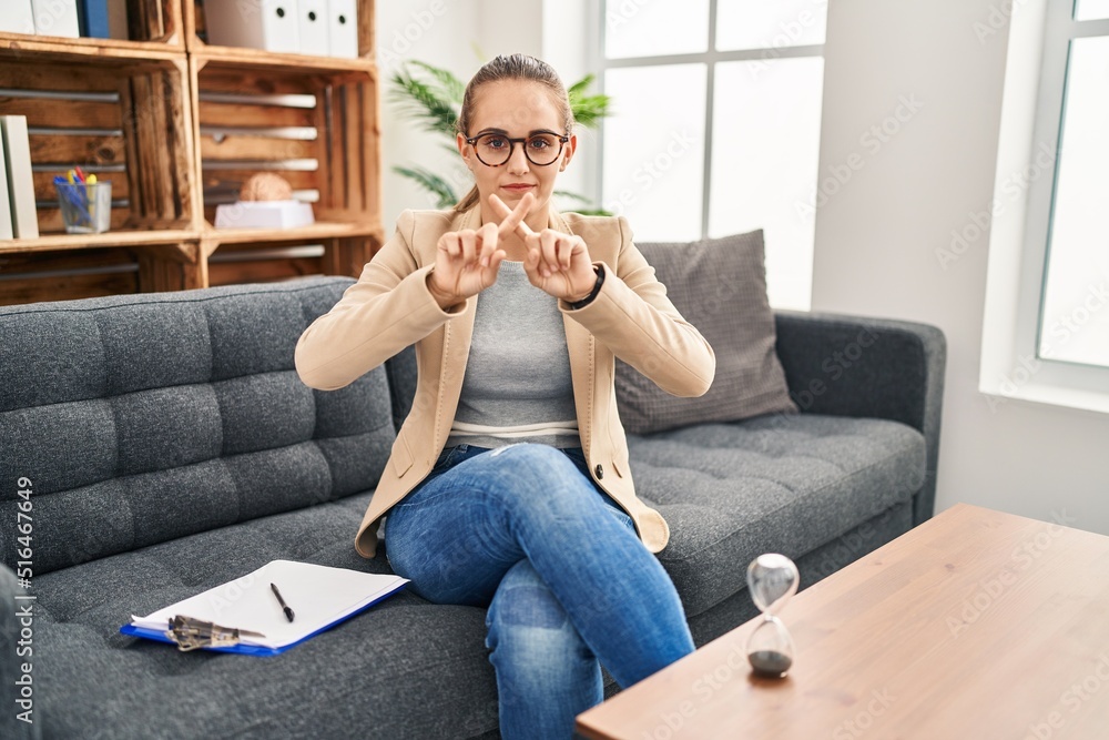 Fototapeta premium Young woman working at consultation office rejection expression crossing fingers doing negative sign