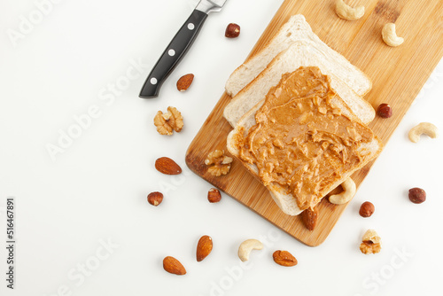 Square bread for toast with peanut butter on a wooden board. Nuts, a knife and a wooden cutting board with a sandwich and slices of bread on a white table.