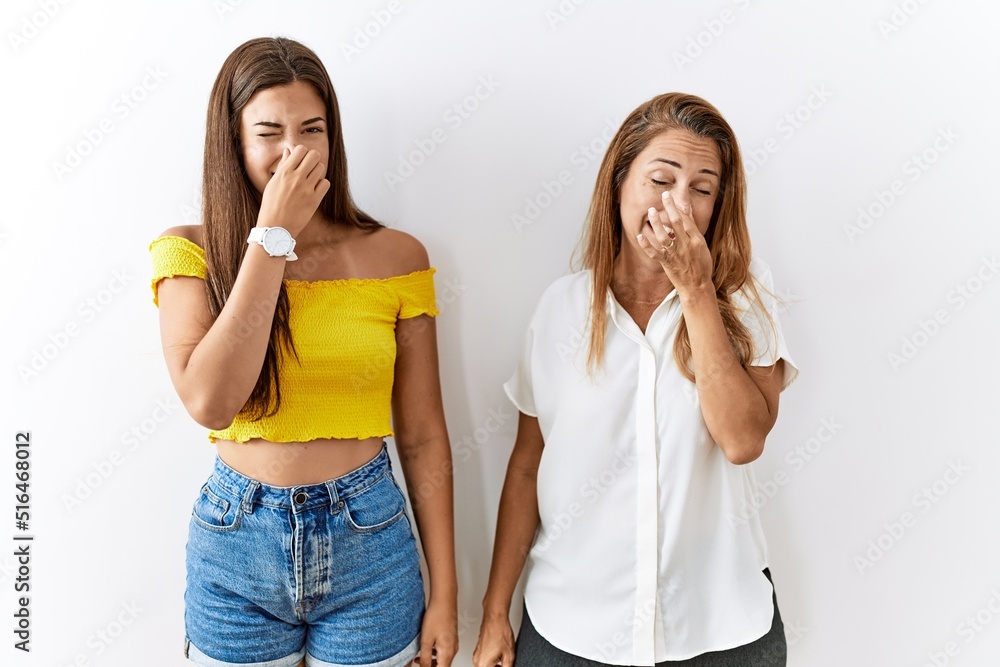 Mother and daughter together standing together over isolated background smelling something stinky and disgusting, intolerable smell, holding breath with fingers on nose. bad smell Mother and daughter together standing together over isolated background smelling something stinky and disgusting, intolerable smell, holding breath with fingers on nose. bad smell