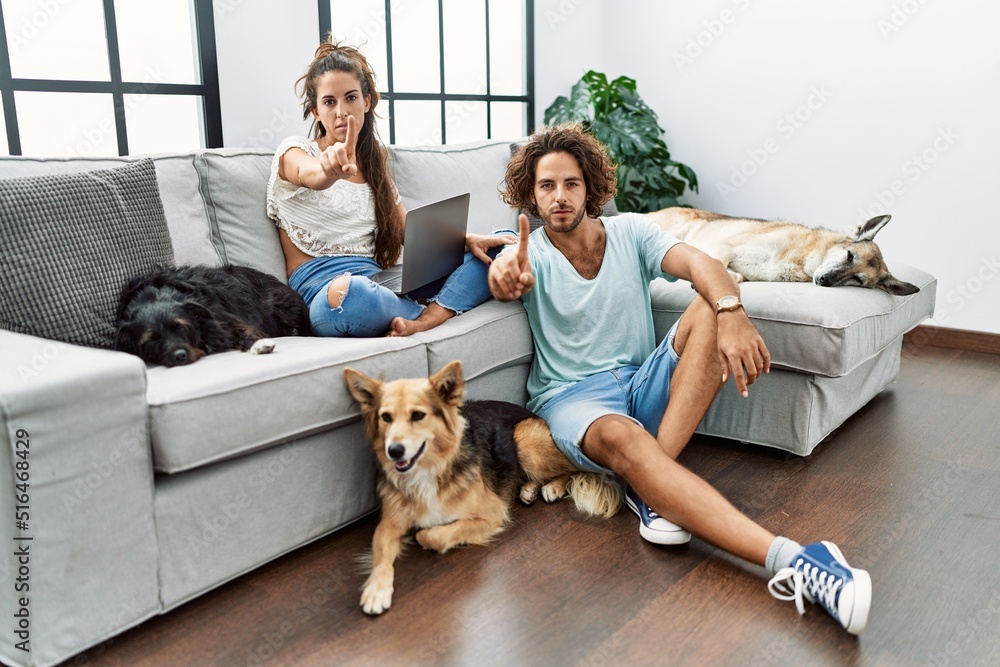 Young hispanic couple with dogs relaxing at home pointing with finger up and angry expression, showing no gesture