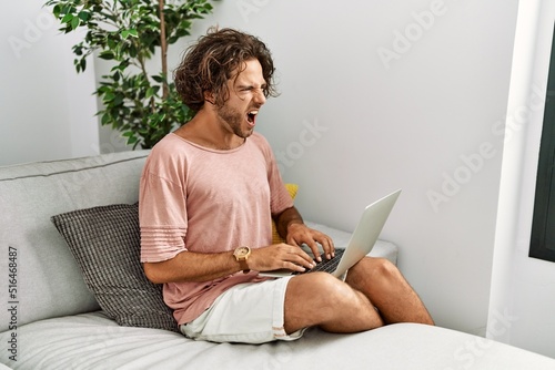 Young hispanic man sitting on the sofa at home using laptop angry and mad screaming frustrated and furious, shouting with anger. rage and aggressive concept.