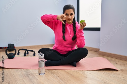 Young hispanic woman sitting on yoga mat eating apple with angry face, negative sign showing dislike with thumbs down, rejection concept