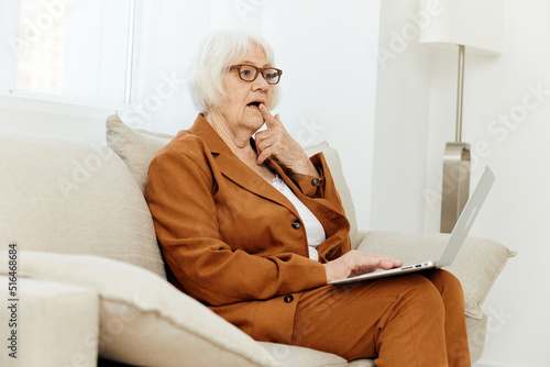 a thoughtful, wise, elderly woman writes an article on a laptop while sitting at home on the sofa in a comfortable environment in a beautiful brown suit and thoughtfully holds her hand near her face