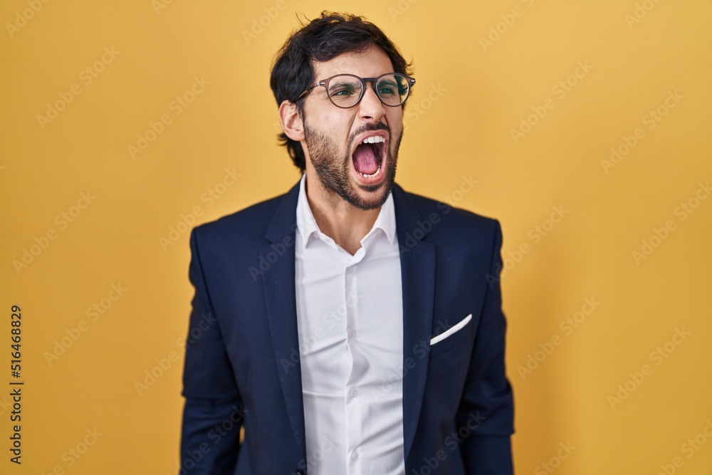 Handsome latin man standing over yellow background angry and mad screaming frustrated and furious, shouting with anger. rage and aggressive concept. Handsome latin man standing over yellow background angry and mad screaming frustrated and furious, shouting with anger. rage and aggressive concept.