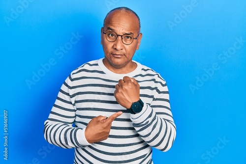 Middle age latin man wearing casual clothes and glasses in hurry pointing to watch time, impatience, looking at the camera with relaxed expression