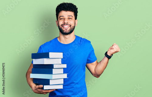 Canvas Print Young arab man with beard holding a pile of books screaming proud, celebrating v