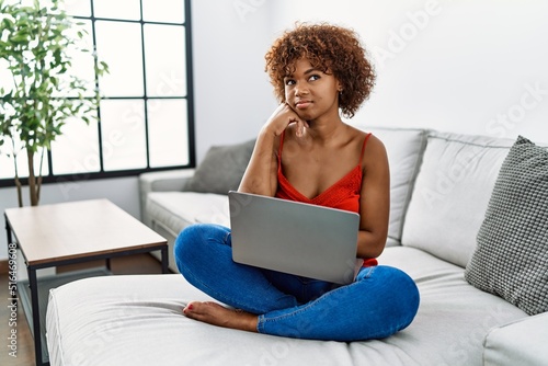 Young african american woman sitting on the sofa at home using laptop with hand on chin thinking about question, pensive expression. smiling with thoughtful face. doubt concept.